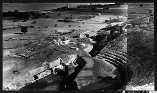 Excavated theatre at Soli with curved stone tiers, low walls and fallen blocks, with fields, a village and the sea beyond