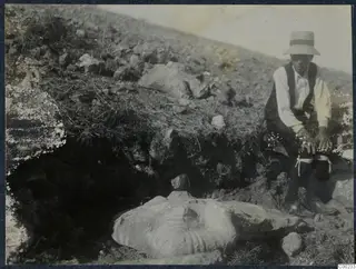Carved stone fragment lying in an excavated hillside at Vouni, with a seated person in a hat beside it