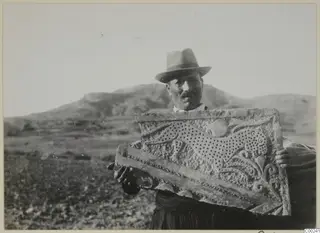 Man in a hat holding a carved stone relief panel with a feline figure and leafy ornament, low hills behind him