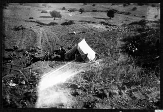 Elevated view of a white tent and seated people on a dry, stony hillside at Soli, with scattered trees.