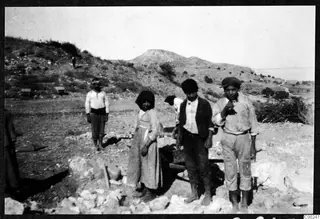 Several people standing among rocks and stones on a dry hillside at Soli, with a wheelbarrow behind them