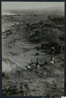 Workers excavating the theatre at Soli on a sloping mound, with wheelbarrows, spoil heaps and a bay in the distance