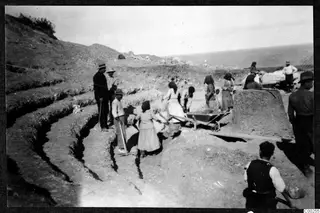 Workers excavating stepped tiers in the hillside at Soli, with women and men shovelling earth beside a wheelbarrow and the sea behind.