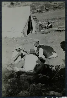 Four men seated around a wooden crate beside a large white tent at a dry, stony camp, with two more people behind.