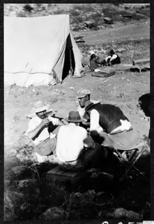 Four men sit around a low wooden box beside a white canvas tent at a stony camp, with two more people behind.