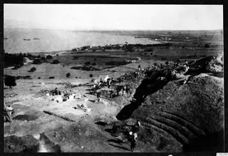 Elevated view of the theatre excavation at Soli, with curved stone tiers, a white tent, workers and the sea beyond