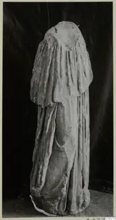 Draped marble sculpture of a standing figure, head missing, photographed against a dark backdrop