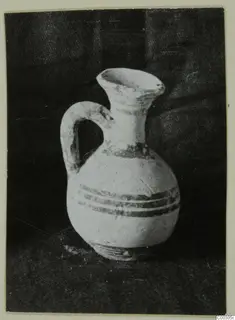 Small white-slipped jug with a loop handle and dark painted bands, photographed against a dark cloth.