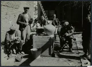 Six men and a child gathered around wooden crates with a large pale jar, in a sunlit courtyard beside a mud-brick wall