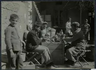 Three men at a wooden crate examining pottery outside a house, with a child and two women nearby