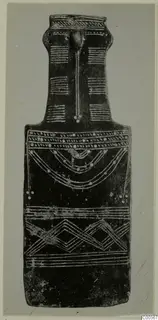 Dark flat idol with a long neck and a raised ridge down the front, decorated with white incised bands, dots, arcs and zigzags