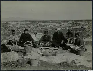 Workers sitting on rocky ground at a picnic at Milia, with plates, baskets and a large jug around them.