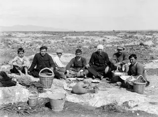 Workers sitting for a picnic on a flat rock at Milia, with baskets, a metal bucket and a large jar beside them.