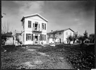 Two-storey white house with shuttered windows and a balcony, with people standing and sitting in the garden in front, Nicosia
