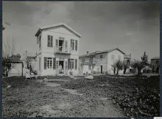 Two-storey white house with a first-floor balcony and people at its doorway, in a garden in Nicosia
