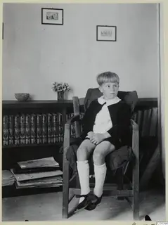 Boy seated in a wooden armchair beside a bookcase of leather-bound books