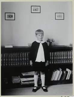 Young boy standing in front of a bookshelf with framed prints on the wall above.