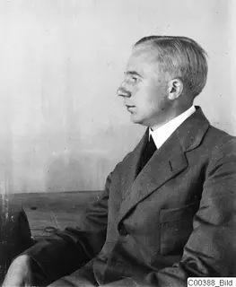Man in a suit seen in profile, seated against a plain pale wall.