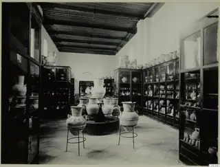 Museum gallery with large painted pottery jars on metal stands and many vessels in glass cabinets