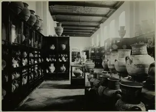 Museum gallery with large painted pottery jars on stands and smaller vessels in glass and wooden cabinets.