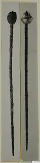 Two long pins with decorated heads, one dark and one lighter, photographed side by side on grey mounts.