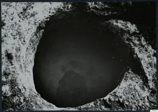 Excavated tomb at Milia seen from above, a round opening in pale soil with a dark mass of stones at the bottom