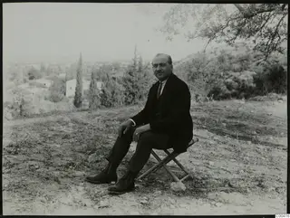 Man in a dark suit seated on a folding stool outdoors, with trees and a white house behind him.