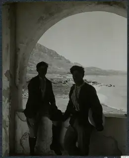 Two men standing in an arched opening, with a bay of sea and a mountain ridge behind them.