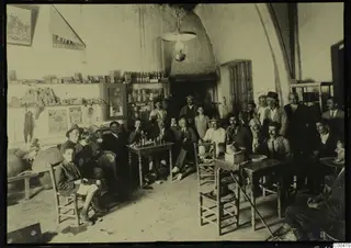 Men seated and standing in a café interior at Lapithos, with bottles on shelves and a hanging lamp overhead
