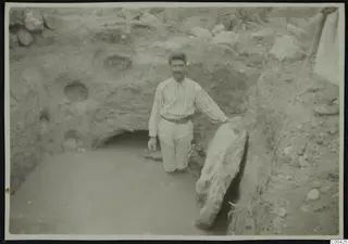 Man standing in water at the bottom of an excavation trench, beside a large pale stone slab
