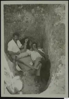 Two men resting in an excavation cutting at Lapithos, with a white helmet on the ground beside them.