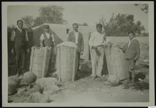 Five men and boys stand beside large wicker baskets and jars in front of a whitewashed building at Lapithos.