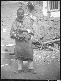 Man with a moustache standing in front of a mud-brick wall, carrying a basket on his back held by a rope.