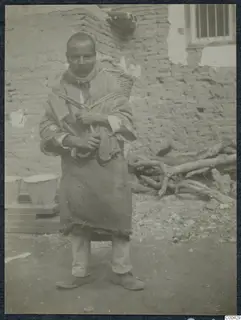 Man with a moustache carrying a basket on his back, standing in front of a mud-brick wall