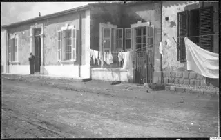 Single-storey plastered house with shuttered windows, a man standing in the doorway and laundry hung on a line in front.