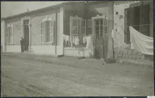Single-storey building with shuttered windows, a standing man at the doorway and laundry hung on a line in front.