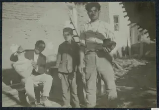 Two men and a boy outside a mud-brick wall; one seated man holds a large pale jar, and a standing man holds a small dark vessel.