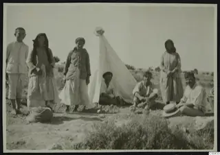 Seven people posed beside a white tent with a straw hat on its peak, on open dry ground