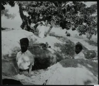 Excavation at Lapithos with two men in trenches and a third man standing beneath a large tree
