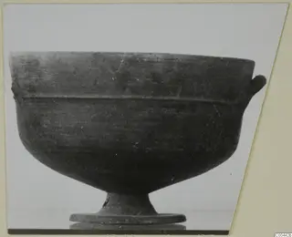 Pottery bowl on a stand, dark surface with a handle-like lug at the right rim and a footed base.