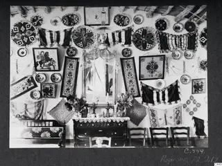 Interior of a house at Milia, a wall hung with embroidered textiles, plates, a mirror and a lamp.