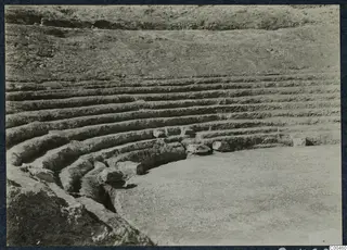 Curved rows of stone seating steps at the theatre at Soli, with a flat paved area below.