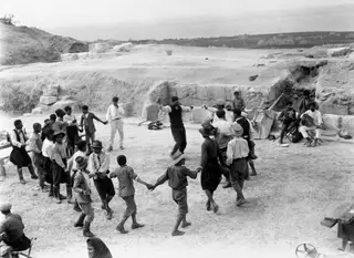 Men and boys dancing in a circle on an excavated area at Soli, with musicians seated at the right beside a stone wall.