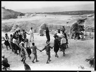 Men and boys dancing in a hand-linked line on the excavated ground of the theatre at Soli, with stone blocks behind.