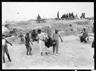 Workers and onlookers at the theatre at Soli, with wheelbarrows and a seated man playing a stringed instrument.