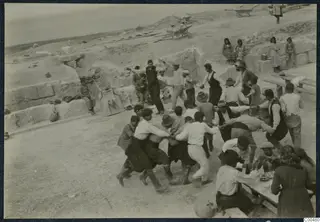 Men dancing in a linked line at the theatre at Soli, with wheelbarrows and onlookers nearby