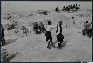 Two men dancing on the bare floor of the theatre at Soli, with seated musicians and workers on wheelbarrows behind them.
