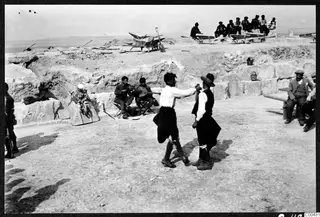 Two men dancing face to face in front of seated musicians at the excavated theatre at Soli.