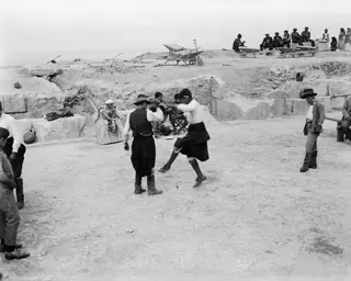 Two men dancing on the open ground of the theatre at Soli, with spectators seated on the stone ledges above