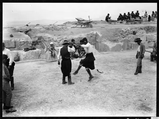 Two men dancing on the bare ground of the theatre at Soli, with onlookers and wheelbarrows on the terrace above